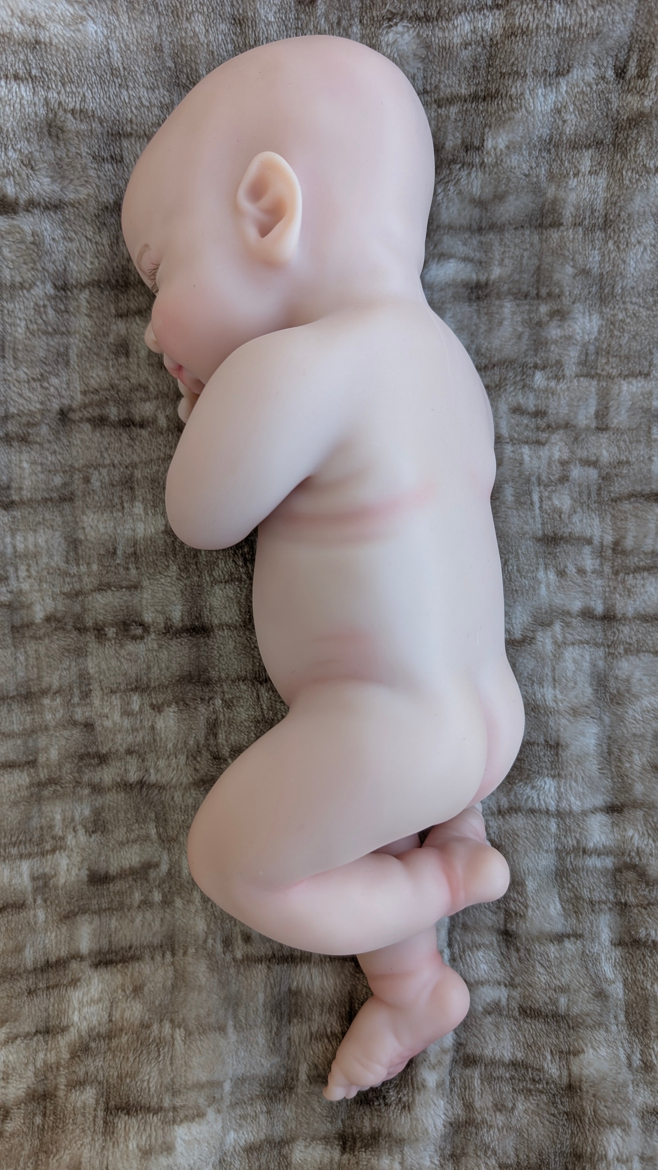 Bebè reborn in silicone di 32 cm con le gambe incrociate in una naturale posizione da neonato - vista laterale